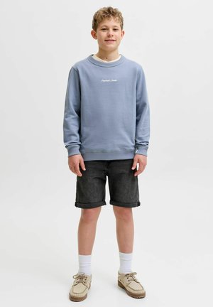 Poiss seisab, seljas hele sinine pusa, mustad üleskeeratud teksapüksid, valged sokid ja beežid kingad, taustal valge lihtne taust.