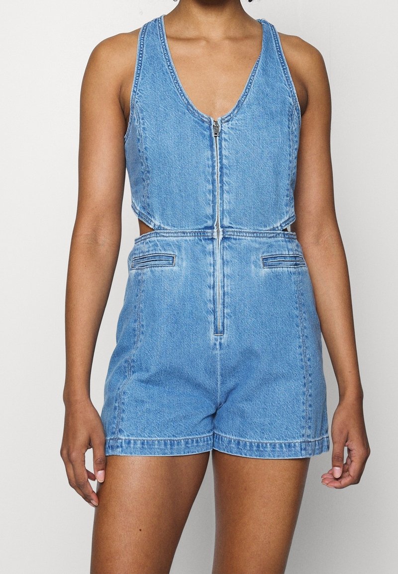 Femme portant une combinaison courte en denim bleu sans manches avec une fermeture éclair à l'avant et des découpes latérales, debout devant un fond clair uni.