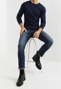Marinblå långärmad skjorta, blå jeans och svarta snörkängor. Modellen sitter på en vit pall mot en ljus bakgrund.