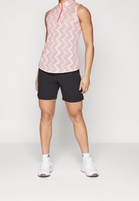Růžový sportovní top bez rukávů se zigzag vzorem a předním zipem. V kombinaci s černými šortkami a bílými sportovními botami.