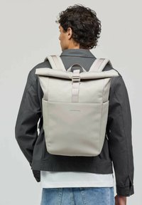 Homme portant un sac à dos beige clair avec une poche avant, vêtu d'une veste sombre et d'un jean bleu clair, tourné le dos à la caméra.