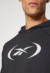Sweatshirt à capuche noire en tissu texturé, avec un cordon de serrage et un grand logo blanc sur la poitrine avec un design circulaire.