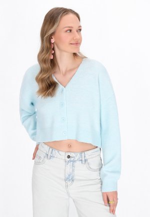 Zugeschnittene hellblaue Strickjacke mit V-Ausschnitt und Knopfleiste; weiche Textur, lange Ärmel, kombiniert mit hochgeschnittenen hellen Jeans.