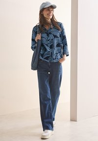Chemise navy avec des motifs à rayures blanches, portée avec un jean en denim foncé et des baskets blanches. Une casquette bleu clair et un sac en denim complètent la tenue.