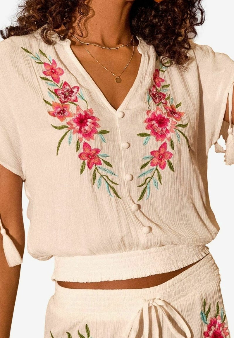 Blouse blanche avec broderie florale en rose et vert, manches courtes avec liens et fermeture à boutons. Tissu texturé, coupe décontractée.