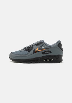 Sneaker atletico grigio con tomaia in mesh e sintetico. Presenta dettagli neri, logo arancione, ammortizzazione Air Max e suola con motivo.