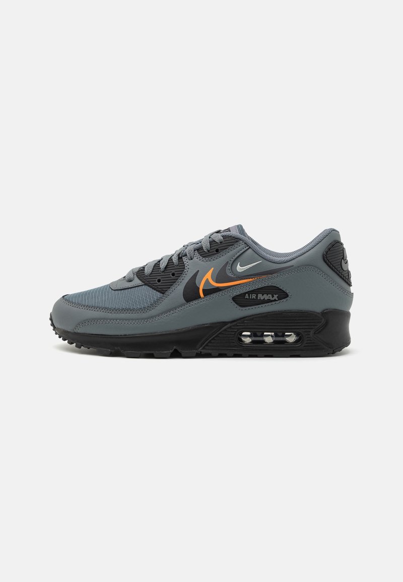 Gray athletic sneaker with a mesh and synthetic upper. Features black accents, orange logo, Air Max cushioning, and a patterned outsole.