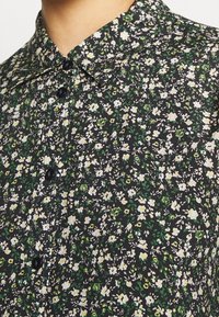 Bluse mit Blumenmuster in Schwarz, verziert mit kleinen weißen und gelben Blumen sowie grünen Blättern, und einem klassischen Kragen.