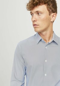 Camicia azzurra chiara con un motivo a trama sottile, maniche lunghe e colletto. Presenta bottoni bianchi lungo la parte anteriore.