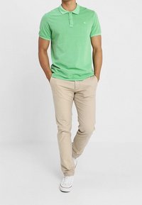 Zielona koszulka polo z krótkim rękawem, z kołnierzykiem i logo, w zestawieniu z beżowymi spodniami. Model nosi białe trampki. Gładka tekstura, luźny krój.