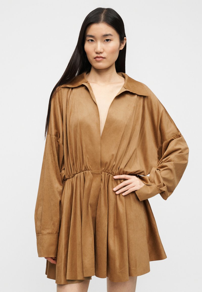 Braunes Wildlederkleid mit tiefem Ausschnitt, langen Ärmeln und geraffter Taille. Fließender A-Linien-Rock mit Falten. Glatte Textur.