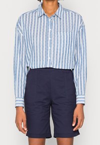 Camicia button-up in cotone a righe blu e bianche con taschino sul petto e maniche lunghe, abbinata a pantaloni corti sartoriali blu navy con risvolti.