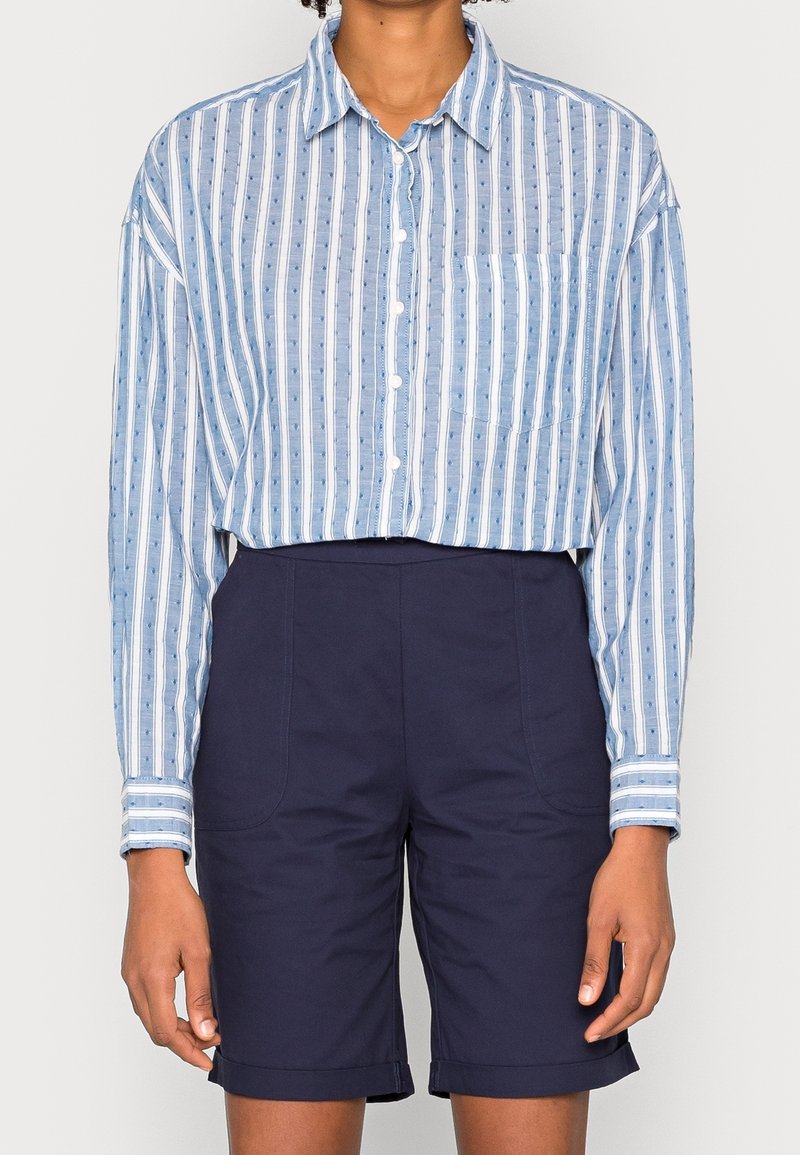 Camicia button-up in cotone a righe blu e bianche con taschino sul petto e maniche lunghe, abbinata a pantaloni corti sartoriali blu navy con risvolti.
