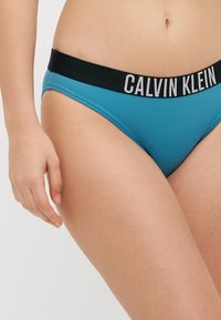 Biquíni em mistura de algodão turquesa com uma faixa elástica preta apresentando o logótipo branco "Calvin Klein". Textura suave, design de cobertura mínima.