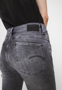 Jeans de denim gris oscuro con un ajuste slim, que presentan un parche de logo de cuero negro, costuras detalladas y un diseño clásico de bolsillos.