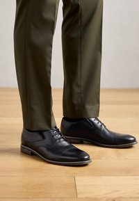 Zapatos de vestir de cuero negro para hombre, con cordones, combinados con pantalones en color verde oliva sobre un suelo de madera.