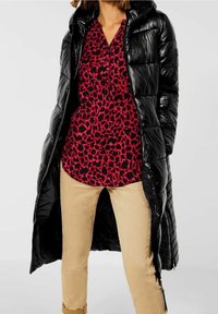 Abrigo largo acolchado negro con un acabado brillante, llevado sobre una blusa con estampado de leopardo rojo y negro, combinado con pantalones beige.