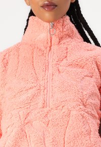 Korallivärinen fleece-pullover korkealla kauluksella, neljännesvetoketju suljenta, ja teksturoitu kangas, jossa on sahalaitakuviota.
