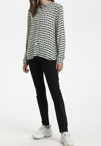 Camicia con bottoni caratterizzata da un motivo zigzag verde, blu marino e crema, maniche lunghe e orlo arrotondato. Abbinata a pantaloni neri aderenti e sneakers bianche.