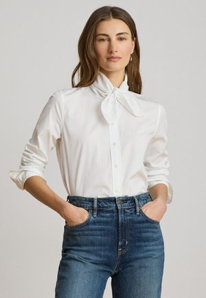 Femme aux longs cheveux bruns, vêtue d'un chemisier blanc avec un nœud au cou et des manches retroussées, associée à un jean bleu taille haute, mains dans les poches.