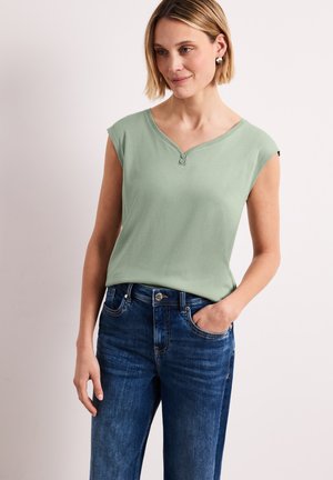 Kobieta w jasnozielonym topie bez rękawów z ozdobnymi guzikami, nosząca ciemnoniebieskie dżinsy, stojąca z jedną ręką w kieszeni.