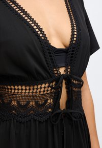 Μαύρο κάλυμμα με βαθύ V-ντεκολτέ, χαρακτηρισμένο από περίπλοκες δαντελένιες λεπτομέρειες και κλείσιμο με κορδόνι στη μέση. Μαλακό ύφασμα με ρευστό σχέδιο.