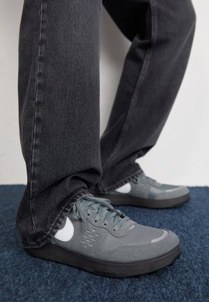 Person wearing gray sneakers with white swoosh logos and dark gray jeans standing on blue carpeted floor.