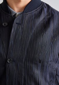 Gros plan d'une veste bleu foncé avec une texture de veinure de bois portée sur une chemise grise à col.