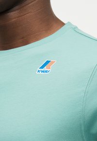 Κοντινό πλάνο του λαιμού και του ώμου ενός ατόμου που φορά ένα ανοιχτό γαλάζιο πουκάμισο με ένα μικρό λογότυπο K-Way σε μπλε και πορτοκαλί στο στήθος.