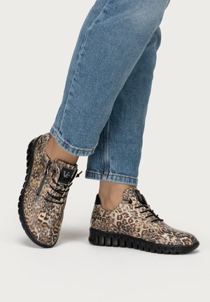 Füße mit leo-geprägten, geschnürten Sneakers mit schwarzen Sohlen, kombiniert mit hochgekrempelten blauen Jeans vor einfarbigem Hintergrund.