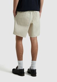 Pantaloni corti in cotone beige con vestibilità comoda, vita elastica e due tasche posteriori. Indossati con scarpe scure e calzini bianchi alla caviglia.