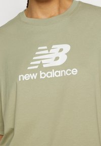Světle olivově zelené tričko, krátké rukávy, vyrobené z měkké bavlny. Na přední straně se nachází výrazné bílé logo "New Balance".