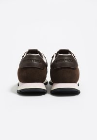 Un par de zapatillas oscuras marrones y blancas vistas desde la parte posterior, con materiales de ante y cuero y con etiquetas de talón de marca.