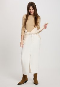 Beige Langarmshirt kombiniert mit einem weißen Jeans-Maxirock mit einem Schlitz vorne. Braune Wildleder-Stiefeletten mit einem ausgestellten Absatz.