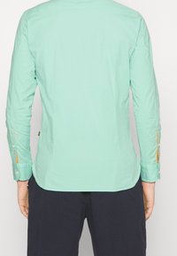 Camicia verde menta a maniche lunghe con polsini a bottone, caratterizzata da dettagli arancioni e un orlo leggermente curvo. Realizzata in materiale di cotone leggero.