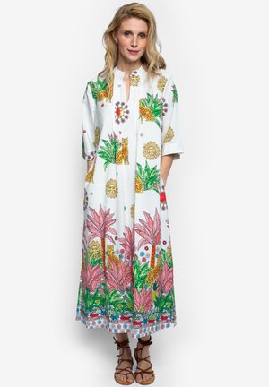 Kobieta ubrana w białą sukienkę sięgającą kostek z kolorowymi tropikalnymi nadrukami roślin, lampartów i słońc, w połączeniu z brązowymi sandałami na sznurowadła.