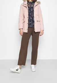 Parka rosa chiaro con cappuccio foderato in pelliccia, indossato sopra un top scuro a fantasia. I pantaloni marroni a costine e le sneakers bianche con dettagli neri completano il look.