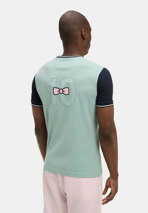 Homme vu de dos portant une chemise vert menthe à manches noires, numéro 10 et motif de nœud rose dans le dos, ainsi qu'un pantalon rose clair.