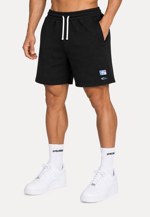 Uomo che indossa pantaloncini neri con coulisse, calzini bianchi con scritto "SMILODOX" e scarpe da ginnastica bianche, in piedi con una mano in tasca.