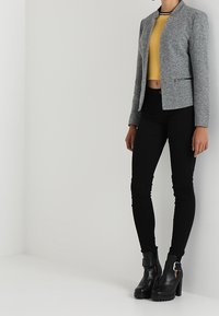 Šedá upravená bunda s kulatým límcem, nošená přes žlutý cropped top; přiléhavé černé kalhoty a černé kotníkové boty na podpatku s přezkami.