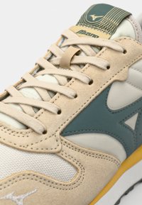 Beige sportcipő, amely velúr és hálós anyagok keverékéből készült, teal színű részletekkel, krémszínű fűzővel és sárga talppal. Figyelemfelkeltő mintás nyelv.