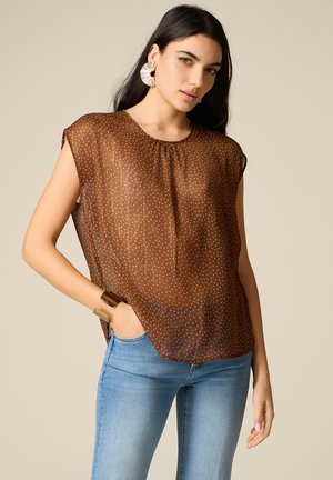 Kobieta o długich ciemnych włosach, ubrana w brązową przezroczystą bluzkę w kropki, niebieskie dżinsy, duże okrągłe kolczyki i dużą bransoletę mankietową.