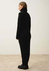 Svart stickad jumpsuit med hög krage, figurnära silhuett och ribbade manschetter. Bärs med svarta snörskor. Slät textur.