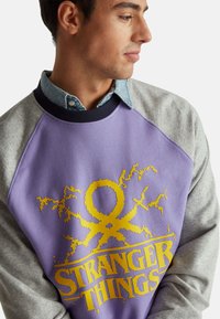 Felpa viola con maniche raglan grigie, caratterizzata da una stampa grafica gialla con il logo e gli elementi di design di "Stranger Things".
