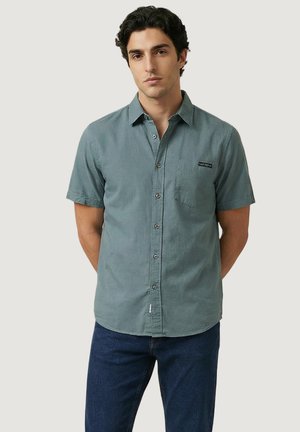 Jonge man met donker krullend haar, gekleed in een teal kort mouw overhemd en donkerblauwe spijkerbroek, staande met handen achter de rug.