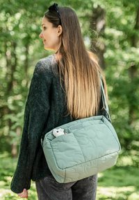 Világos zöld, steppelt válltáska, texturált mintázattal, amelyből egy kis plüssjáték kandikál ki a zsebből. Az állítható pánt látható.