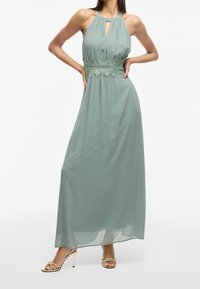 Maxiklänning i mjukt grönt tyg med halterneck och nyckelhålsdetalj. Har spetskant vid midjan, flytande design och slät textur.