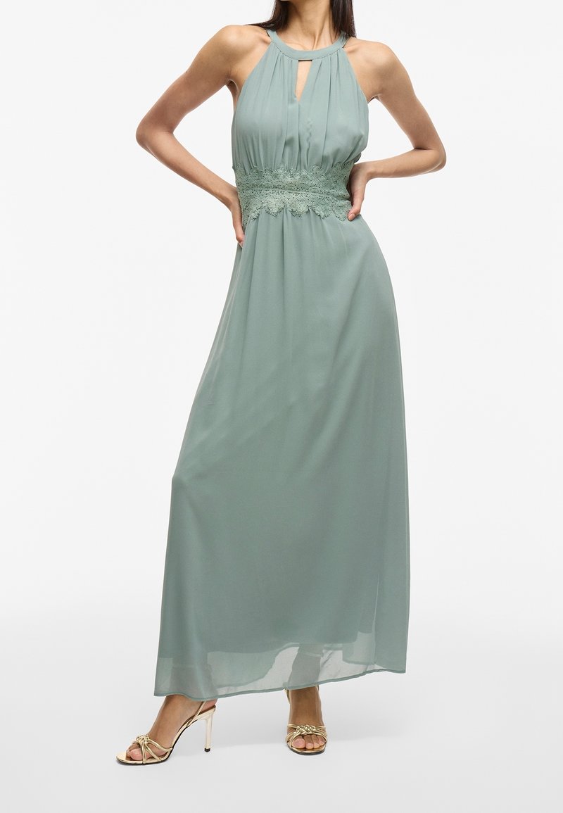 Maxiklänning i mjukt grönt tyg med halterneck och nyckelhålsdetalj. Har spetskant vid midjan, flytande design och slät textur.