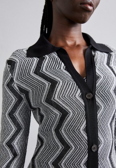 Juodai balta zigzag rašto kardiganas su apykakle, mygtukų uždarymu ir tekstūruotu audiniu, turinčiu metalinių akcentų.