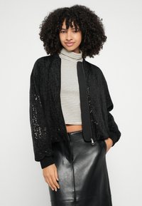 Juoda bombonešio striukė su blizgučių dekoracijomis, raukšlėtais rankogaliais ir užtrauktuku priekyje, derinama su dryžuotu aukštu kaklu ir dirbtinės odos sijonu.